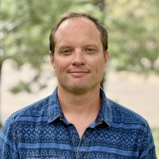 Ian Cooley, smiling in a blue patterned shirt in front of green trees
